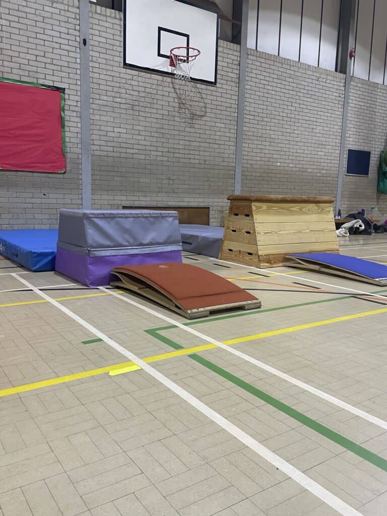Balancing equipment and mats set up on a gymnasium floor, including a vaulting box, foam blocks, and soft landing mats, with a basketball hoop and gym wall in the background.