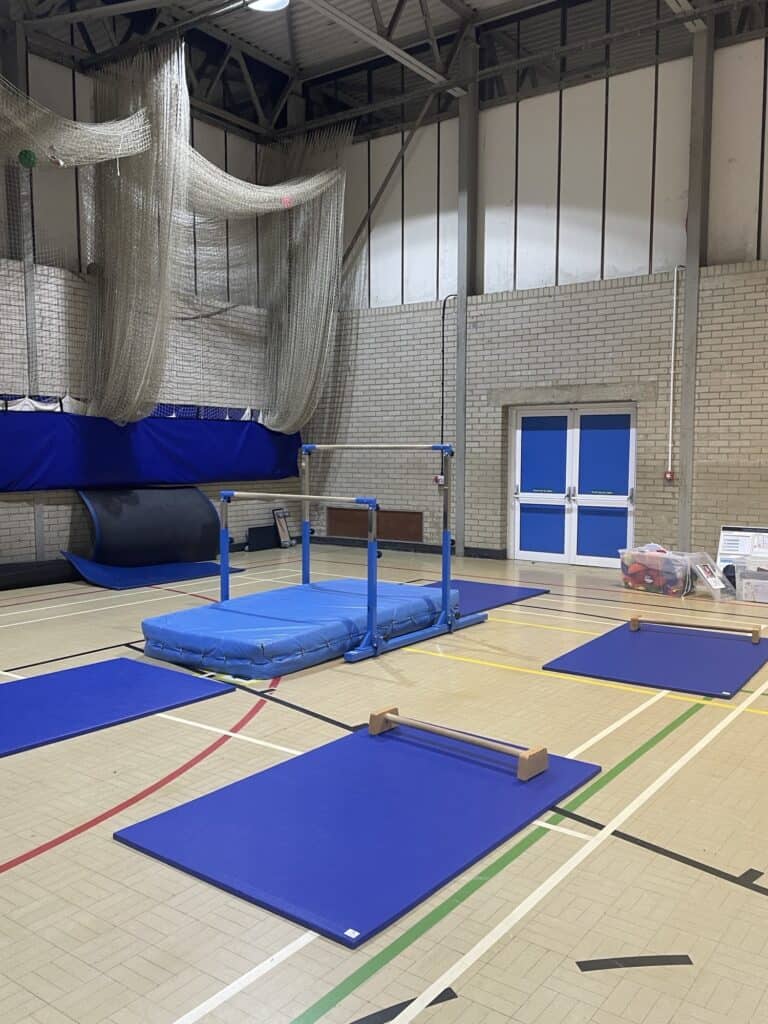 Blue gymnastics mats and vault equipment in an indoor sports hall with climbing nets on the wall.