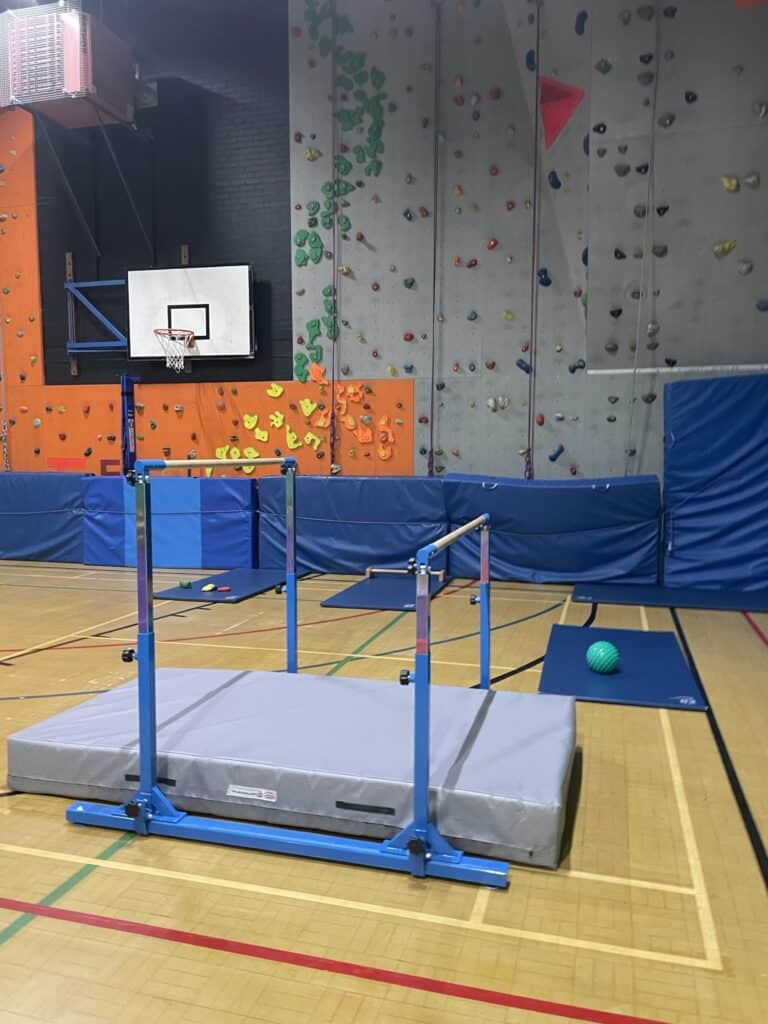 Climbing wall and sports equipment set up in an indoor gym with a basketball hoop in the background. The gym features colourful climbing holds and padded safety mats on the floor.