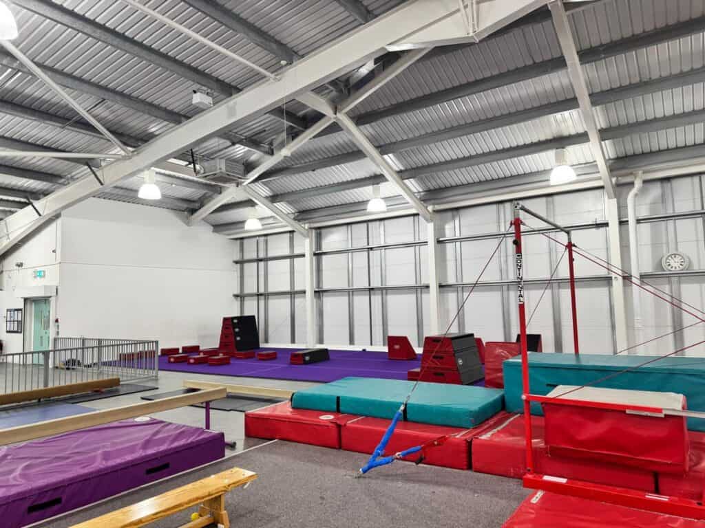 An indoor gymnastics training facility with various equipment including mats, bars, and climbing structures under a high, metal roof.