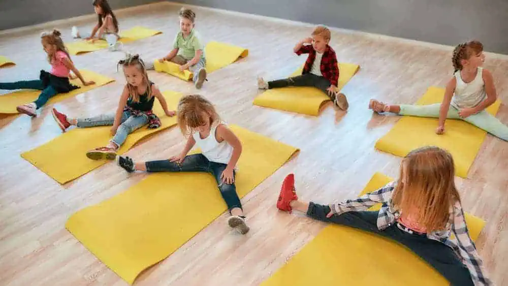 Children participating in a yoga class, performing leg stretches on yellow mats in a bright, spacious room. Kids are engaging in a fun, relaxing activity designed for flexibility and relaxation.