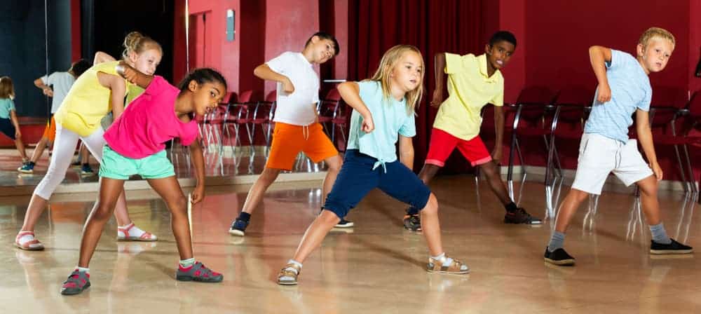 Kids dance class indoors, children energetic dance movements, diverse group of kids in colourful casual clothing, active kids enjoying dance class, children having fun dancing together in a studio.