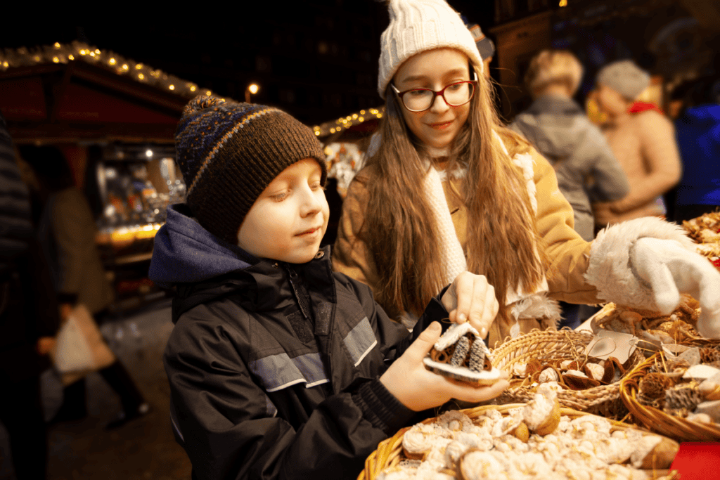 Kids shopping for festive treats at a Christmas market in the evening, surrounded by holiday lights and decorations, enjoying the seasonal atmosphere.