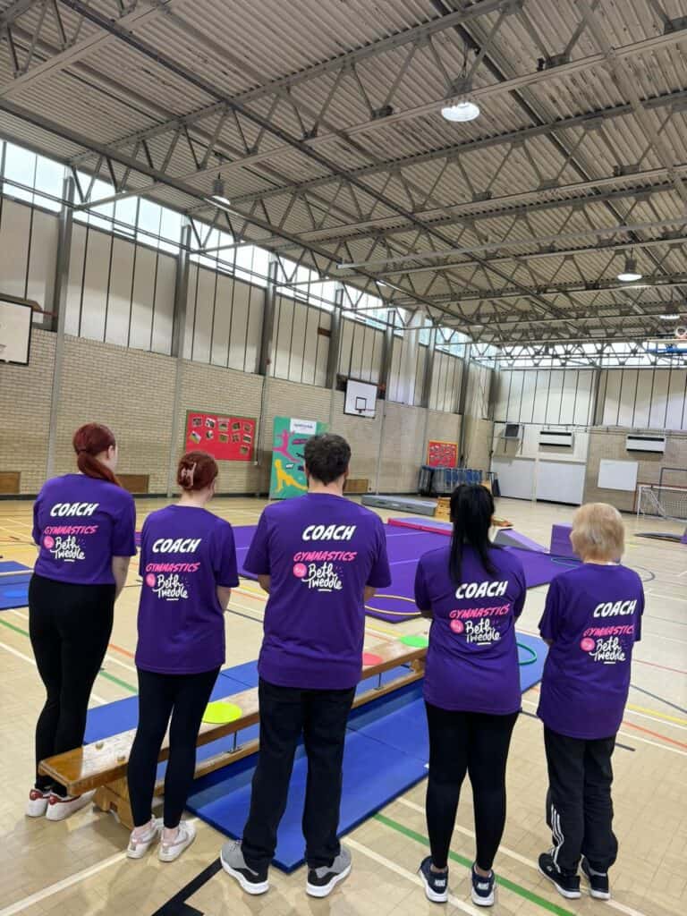 Coaching staff standing in a gymnasium during a gymnastics training session, wearing purple t-shirts with "COACH GYMNASTICS by Betweddle" printed on the back, with various gymnastic equipment set up around.