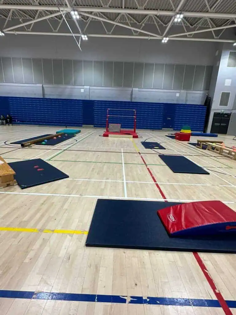 Flexible gymnastics and physical activity equipment set up in an indoor sports hall for children’s exercise and play. Equipment includes mats, balance beams, climbing blocks, and gymnastics bars for kids' activities.