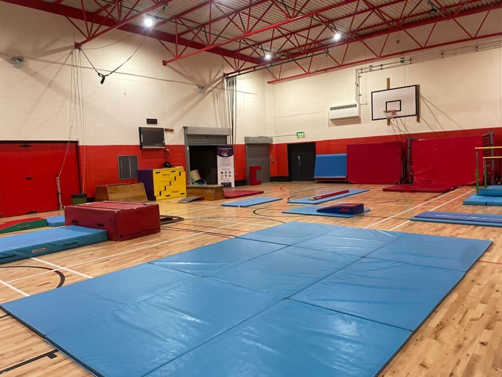 Clear gymnasium with gymnastics mats, vault boxes, and climbing equipment set up on a wooden sports hall floor. Red and white walls with sports facilities like basketball boards and padded safety barriers.