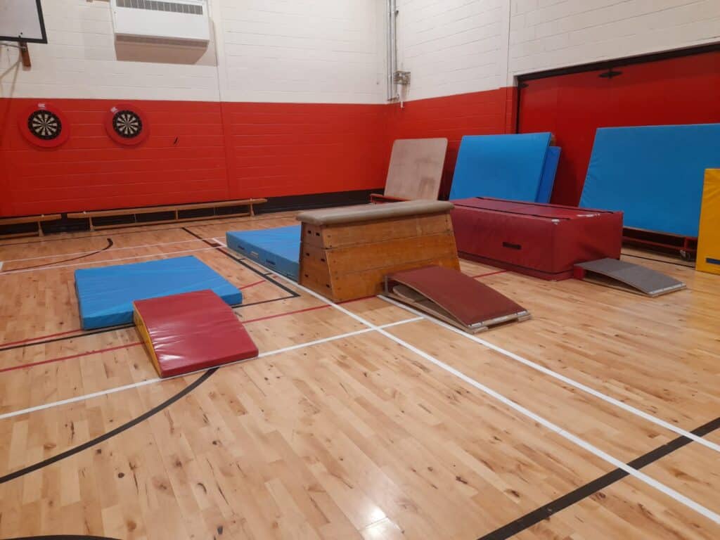 Climbing and gymnastic equipment set up on an indoor sports hall wooden floor, including mats, vaulting box, and soft landing pads, ready for training or gymnastics practice.