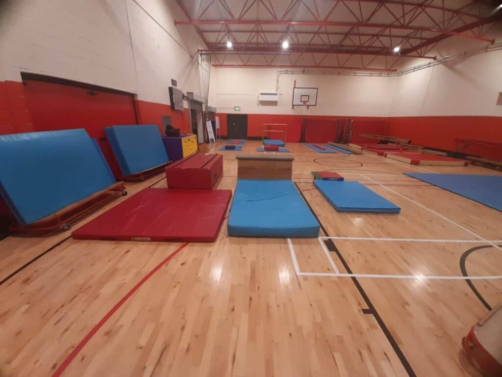 Colourful gym mats and equipment arranged in a spacious indoor sports hall with a wooden floor and high ceiling. The hall is set up for gymnastics training or activities, with padded mats and sliding tables.
