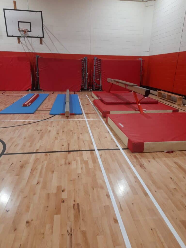 High jump and gymnastics equipment set up in an indoor sports hall with a wooden floor, padded mats, and wall-mounted basketball hoop for athletic training.