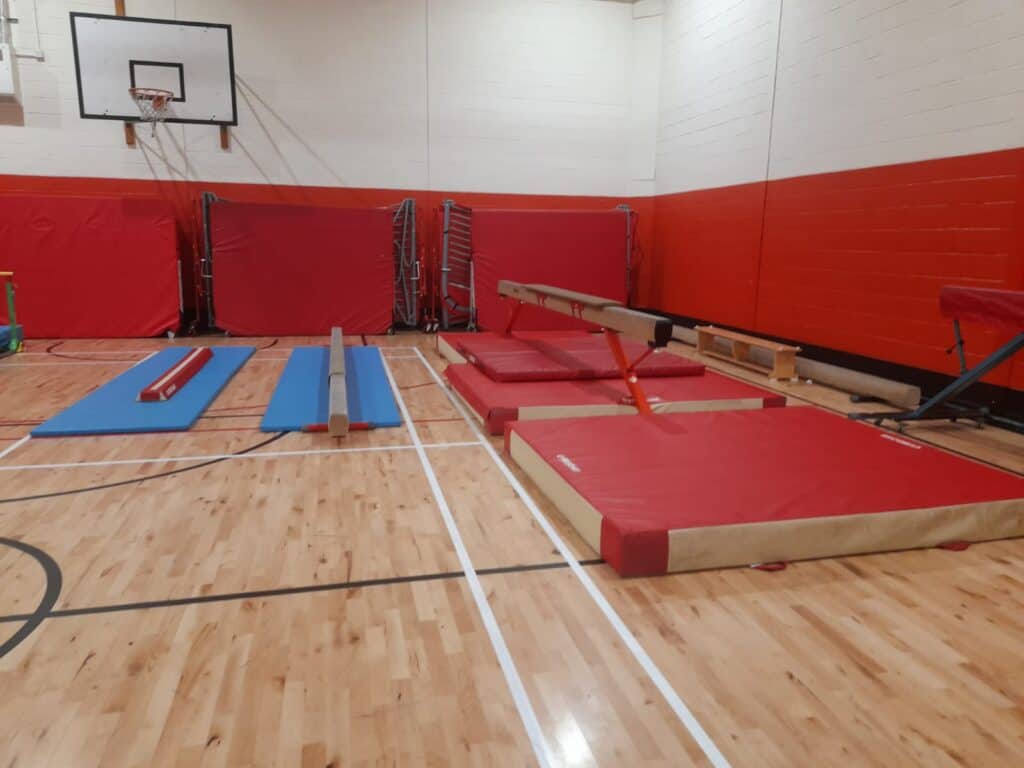 A gymnastics training area in a gymnasium featuring mats, a vaulting table, and training apparatuses on a wooden floor with red wall padding.