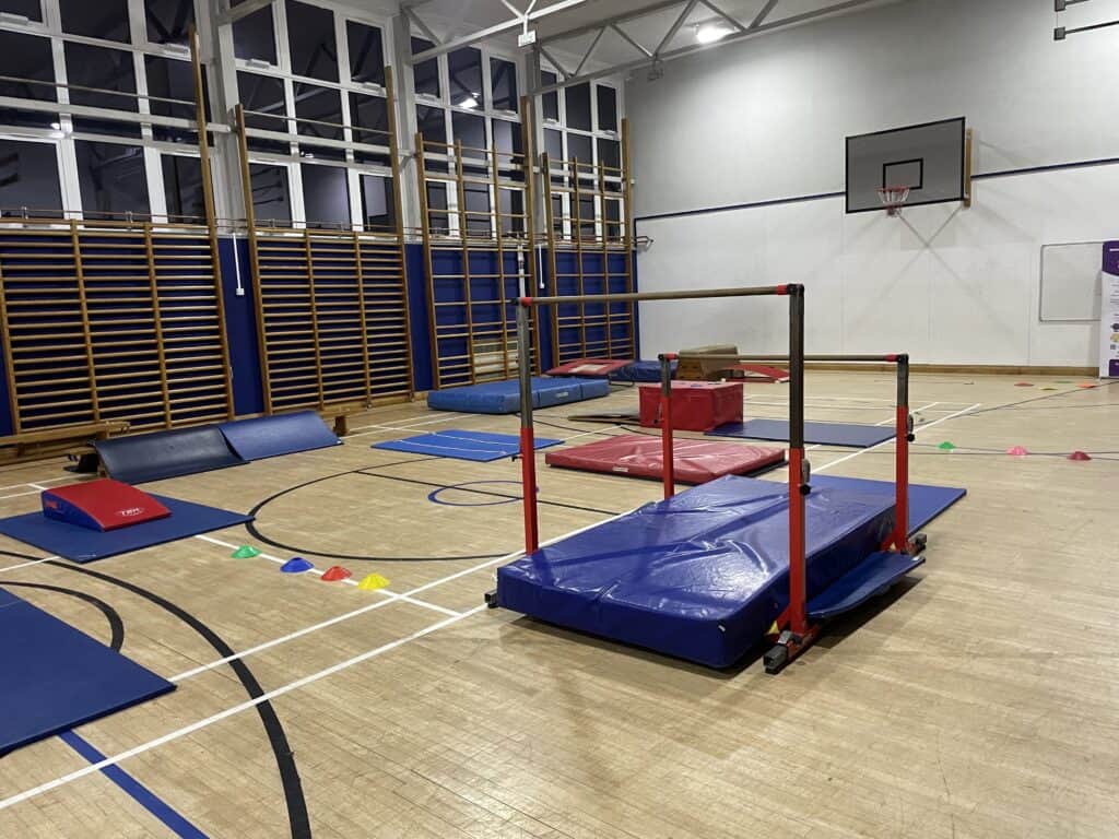Climbing and gymnastic equipment setup in a spacious indoor sports hall for children's activities, including mats, bars, and cones for obstacle courses.