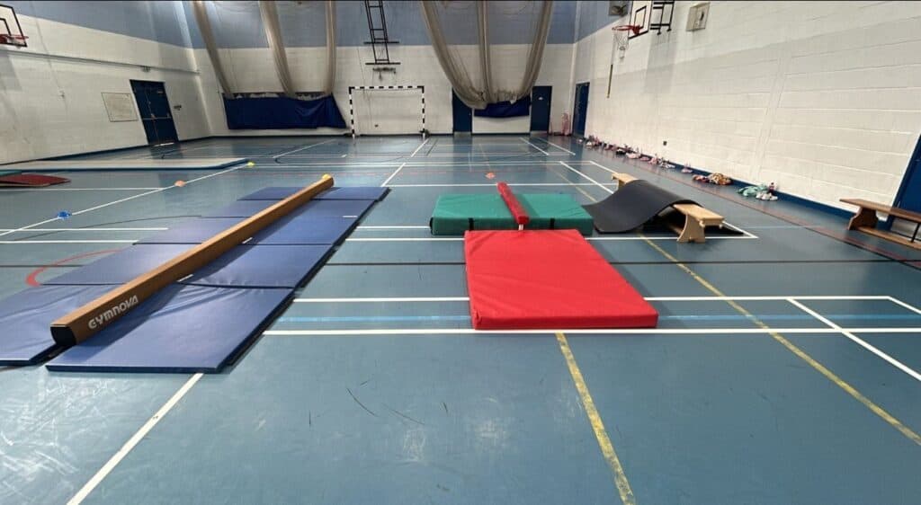 Gymnastics training area with mats, vaults, and a mini trampoline in an indoor sports hall. Equipment is set up for gymnastics practice or physical education activities.