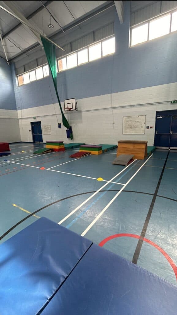 A gymnasium with various gymnastics equipment including mats, vaulting tables, and a climbing rope, set up for indoor physical activities or training sessions. The space features high ceilings, large windows, and a polished sports floor.