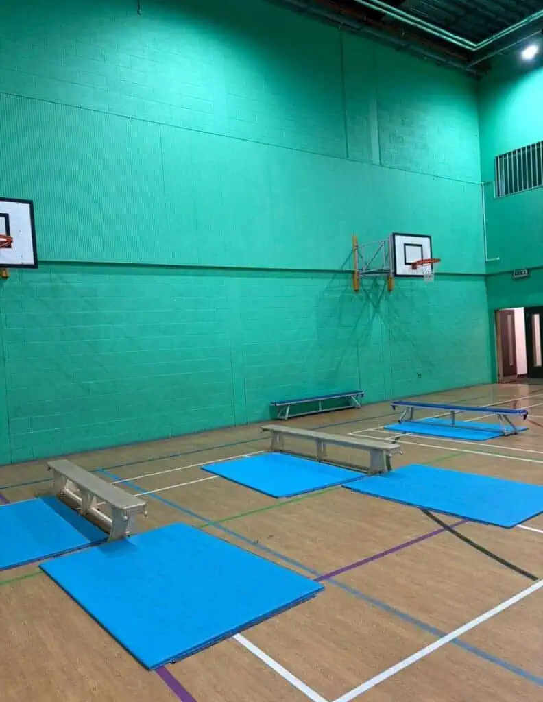 An empty indoor gymnasium with green walls, two basketball hoops, and blue gym mats laid out on the wooden floor for exercise or training activities.
