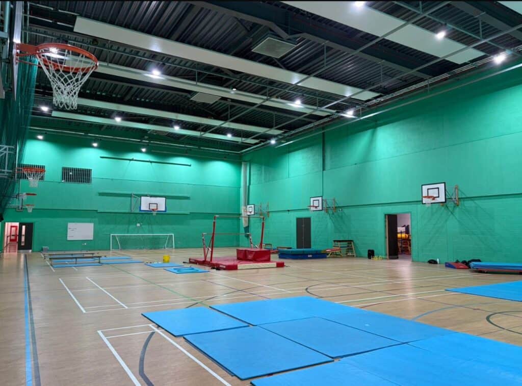 Indoor sports hall with basketball hoops, gym mats, and training equipment in a spacious, well-lit gymnasium with green walls and wooden flooring.