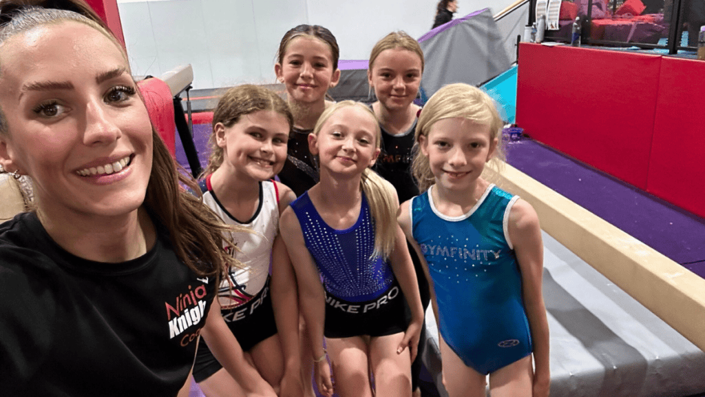 Cheerleading team group photo of young girls in gymnastics outfits smiling at an indoor sports facility.
