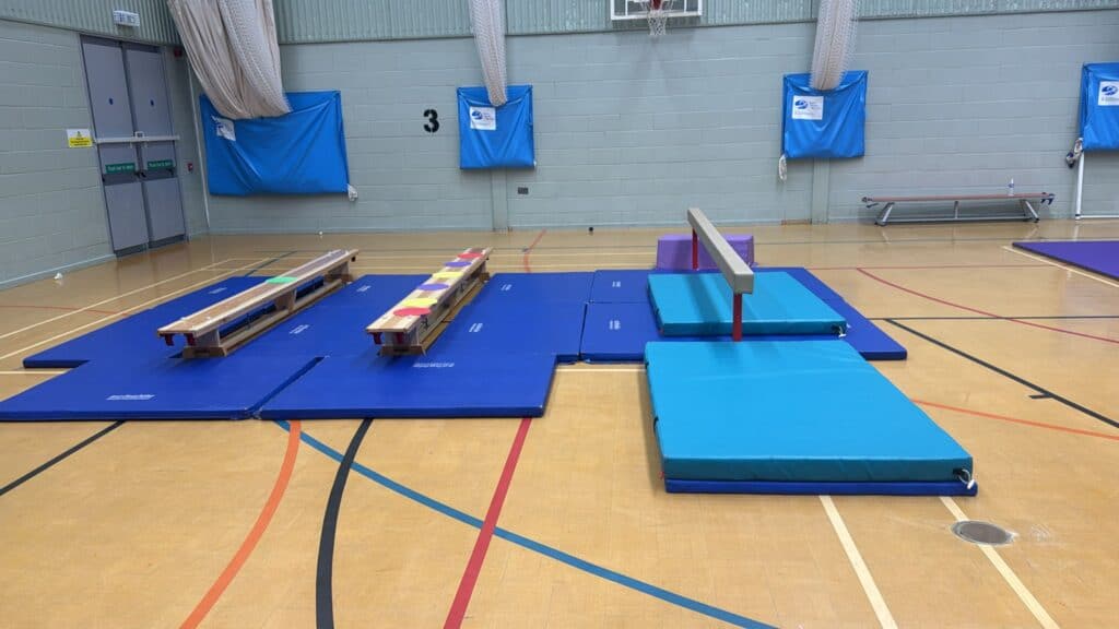 Blue gym mats, benches, and equipment set up in a sports hall for gymnastics or physical training activities. The area is inside a large indoor gymnasium with a wooden floor.