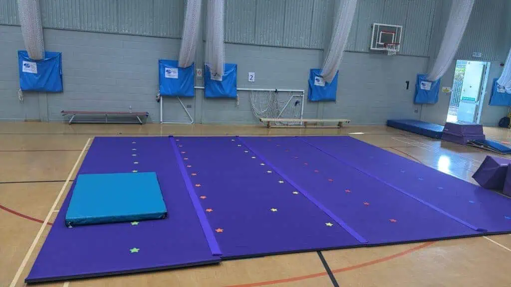 Bright purple gymnastics mats with colourful star-shaped stickers arranged in lines on a wooden sports hall floor, ready for training or competitions.