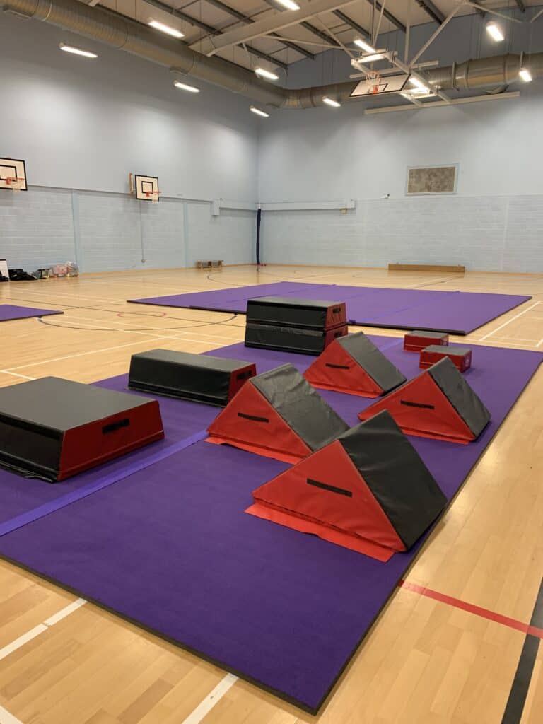 Red and black gymnastic mats and training equipment on purple mats inside a spacious sports hall.