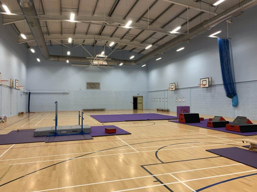 Spacious indoor gymnasium with wooden flooring, equipped with various gymnastics mats and equipment, suitable for training and physical education activities. The room has high ceilings, good lighting, and wall-mounted basketball hoops.