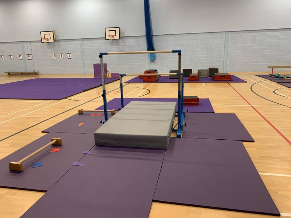 Flexible gymnastic equipment setup on purple mats in a sports hall, including a vault, parallel bars, and stepping boxes for gymnastics training and practice.