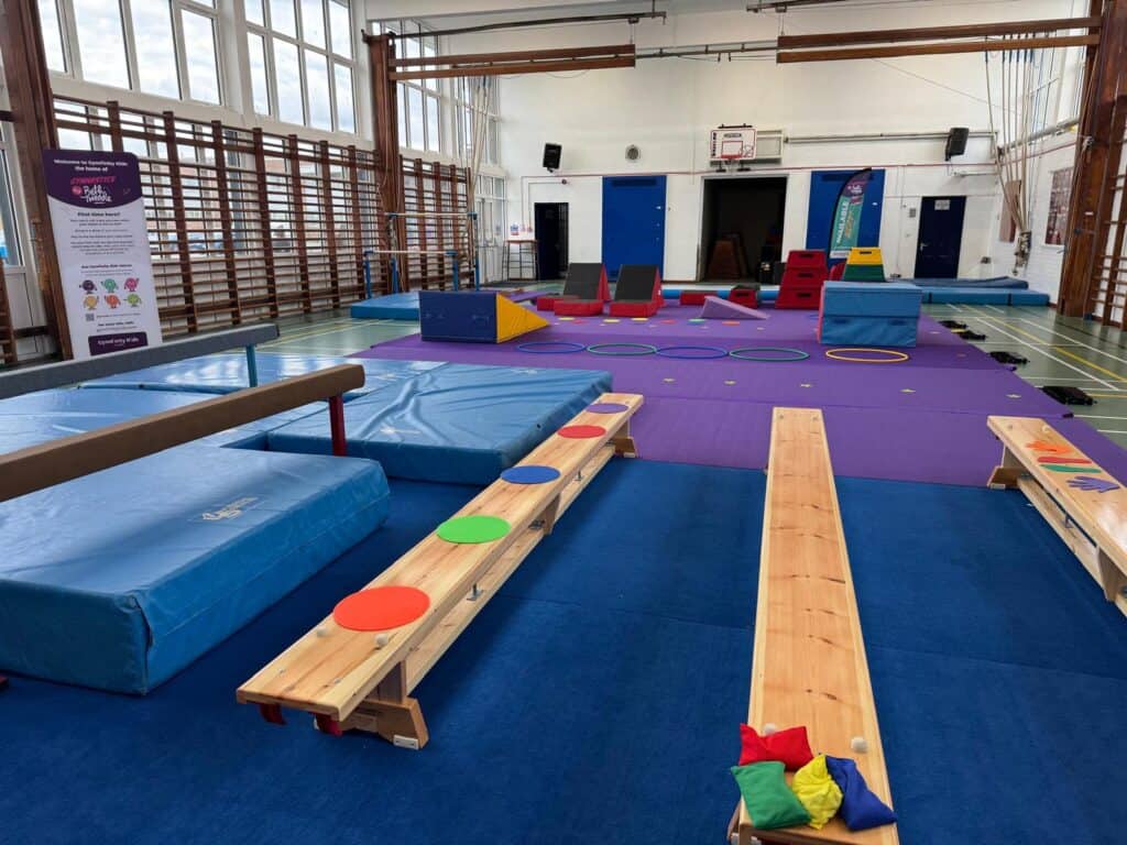 A colourful indoor gymnasium setup for children's activities with foam mats, wooden obstacle benches, hoops, and soft play equipment in a spacious sports hall.
