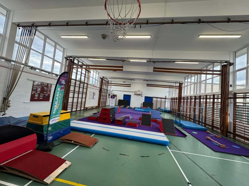 Gymnastics training area with various equipment, mats, and a basketball hoop in a spacious indoor sports hall illuminated by natural light from large windows.