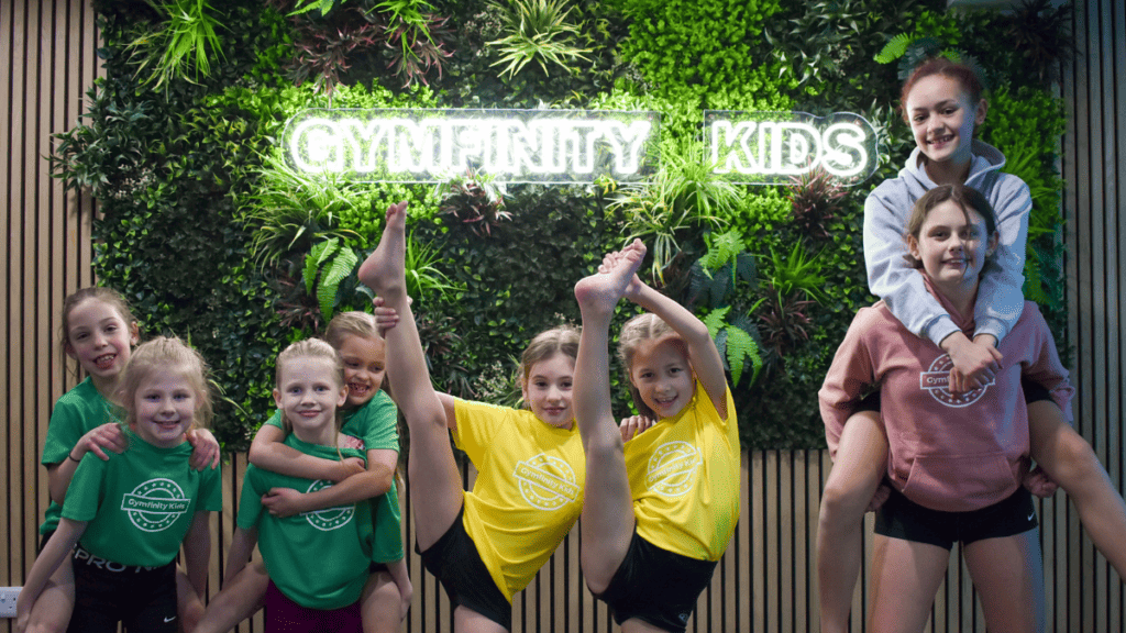 Gymfinity kids children performing indoor gymnastics in a fun, vibrant environment with a green wall background and illuminated "GYMFINITY KIDS" sign.