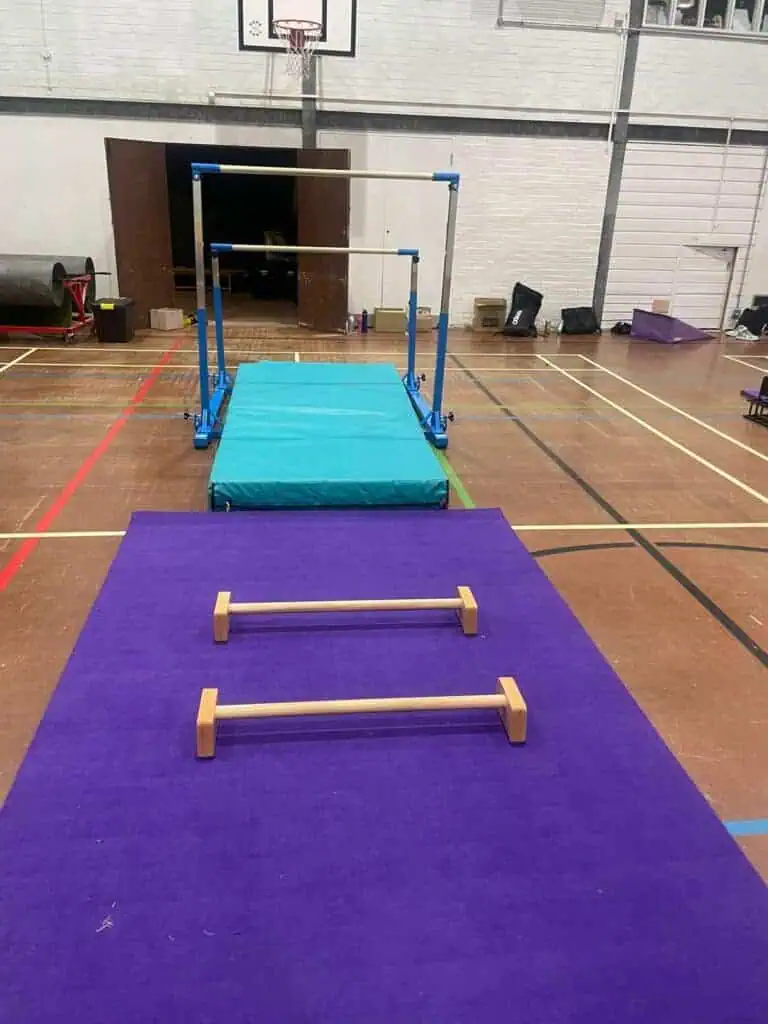 Fast gymnastic training area with padded mats, parallel bars, and agility hurdle setup inside a sports hall. The space is prepared for gymnasts to practice routines and improve skills.