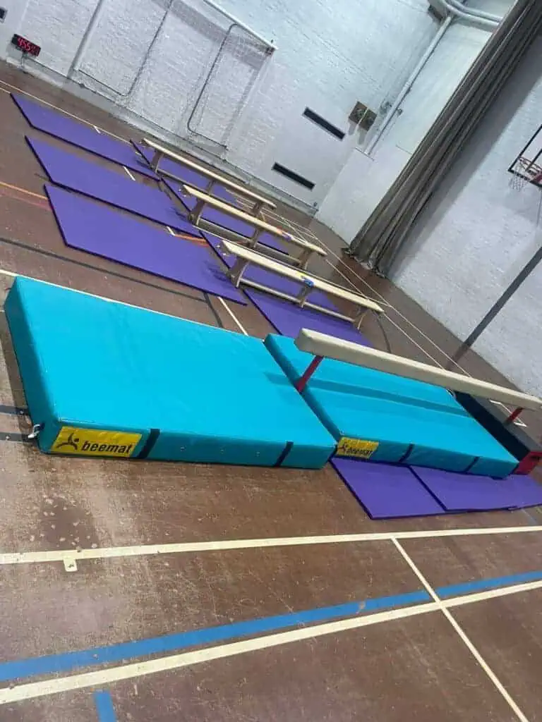 Portable gymnastics mats and vaulting horse set up on a gymnasium floor, ready for training or competition. The equipment includes blue cushioned mats, vaulting boxes, and a vaulting table for gymnasts.
