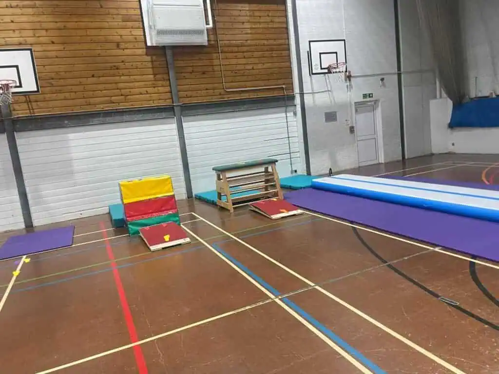 Colourful gymnastic mats and climbing equipment set up in an indoor sports hall with basketball hoops and wooden and metal walls.