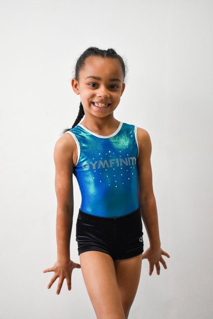 Gymnast girl in a blue leotard smiling confidently after a performance or practice session.
