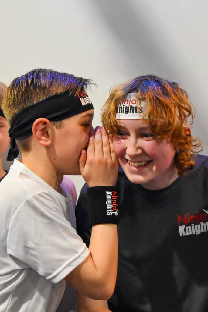 Excited young children sharing a secret at Ninja Knights competition, wearing team headbands and wristbands, displaying team spirit and camaraderie.
