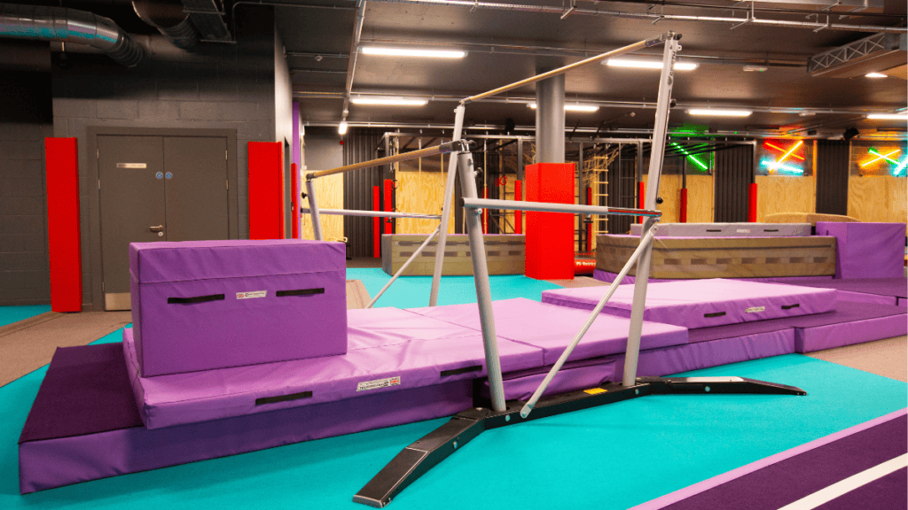 Soft-play gymnastics equipment in an indoor children's play area, with purple padded mats and a balance beam on a vibrant blue and purple floor.