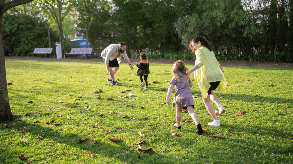 Playing children and parents enjoying a sunny day in a park, with kids running across the grass and leaves, parents supervising and having fun together, surrounded by trees and benches in the background.