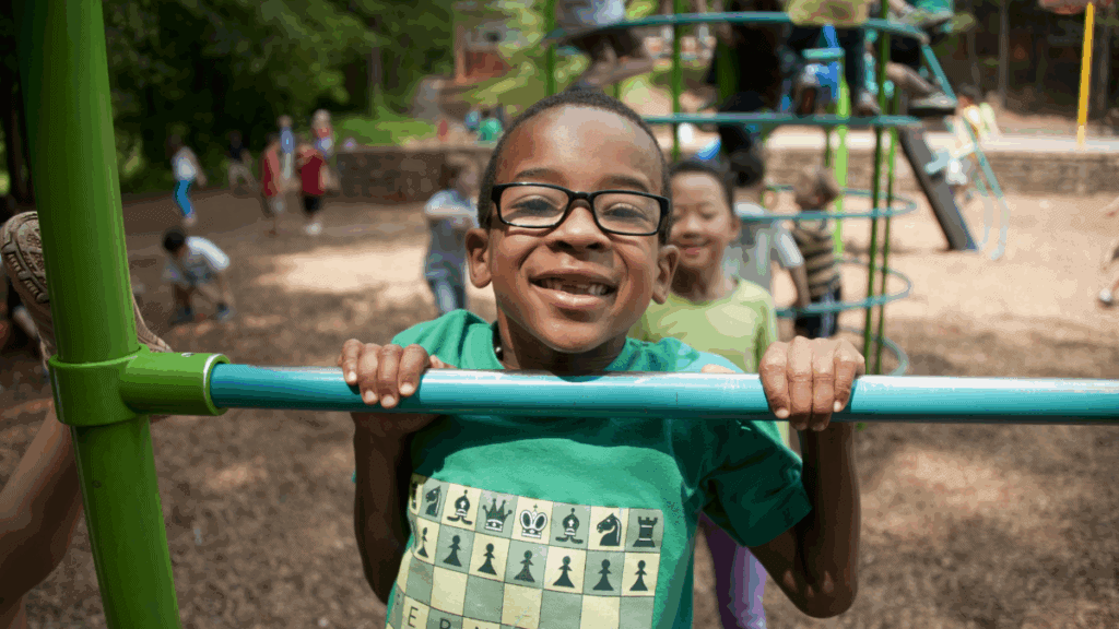 Children playing on a jungle gym at a playground, smiling and enjoying outdoor activity. A young boy in glasses is holding onto the bars, with other kids visible in the background. The setting is a lively, green park.