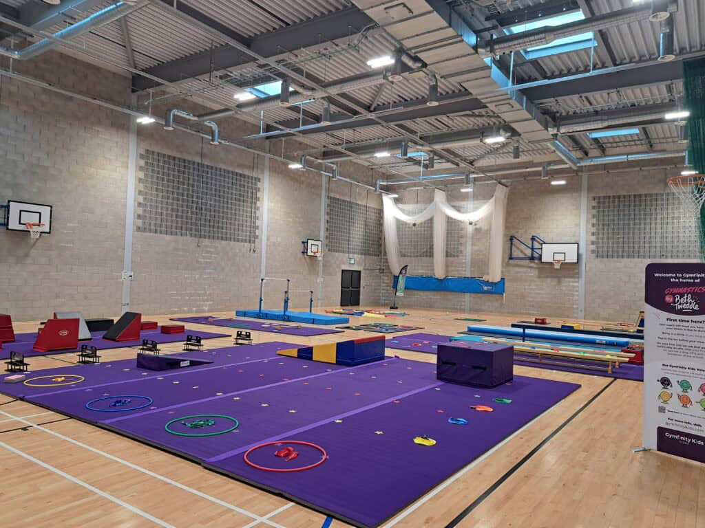 A spacious indoor gymnastics training area with various gymnastic equipment including mats, rings, and bars in a well-lit sports hall with a high ceiling and brick walls.