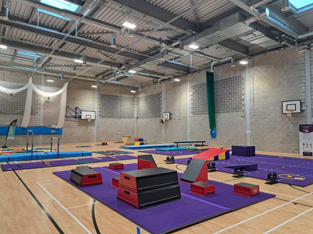 A detailed gymnasium setup with various training equipment including mats, balance beams, and obstacle courses on a purple floor. The space is well-lit with high ceilings and sports facilities.