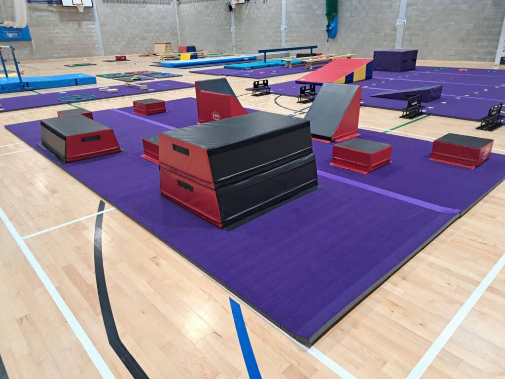 Padded gymnastics training area with various mats, vaults, and obstacle equipment, set up indoors on a wooden sports hall floor for gymnastics practice or events.