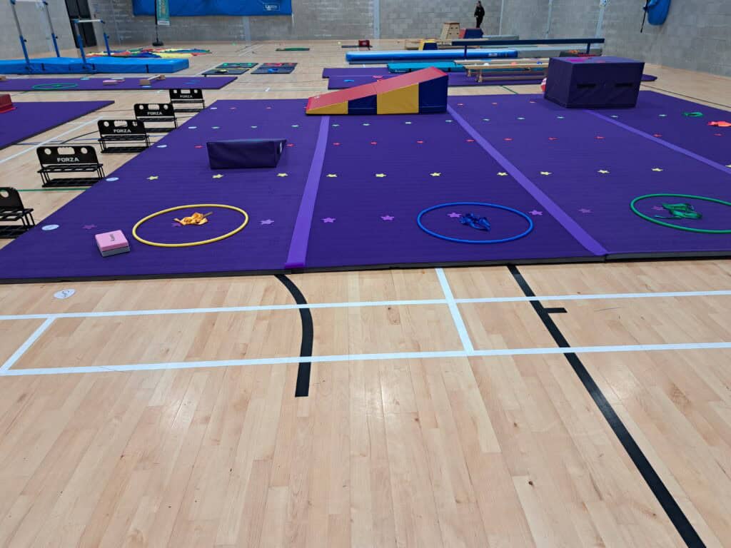 Colourful indoor gymnastics setup with mats, hoops, and foam blocks for training exercises. The gym has purple mats, bright equipment, and a spacious area for various gymnastics activities and training sessions.