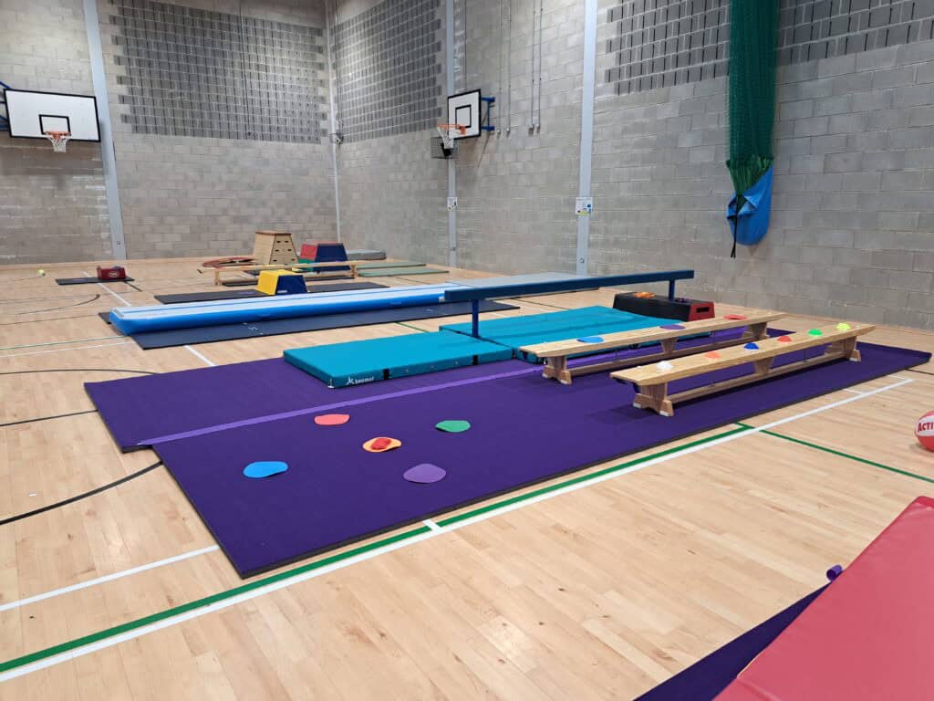 Climbing frames, mats, and stools set up in an indoor sports hall for gymnastics or physical activity training. The hall features a wooden floor, high walls, and basketball hoops.