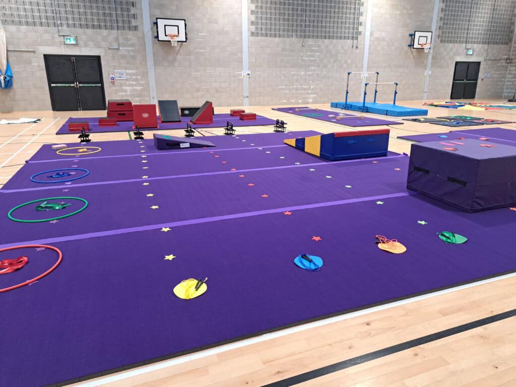 Colourful gymnastics equipment set up on purple mats inside a sports hall with basketball hoops and brick walls.
