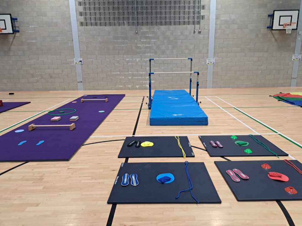 Climbing and gymnastic equipment set up in a sports hall for training or fitness activities with safety mats, hurdles, ropes, and colourful slings.