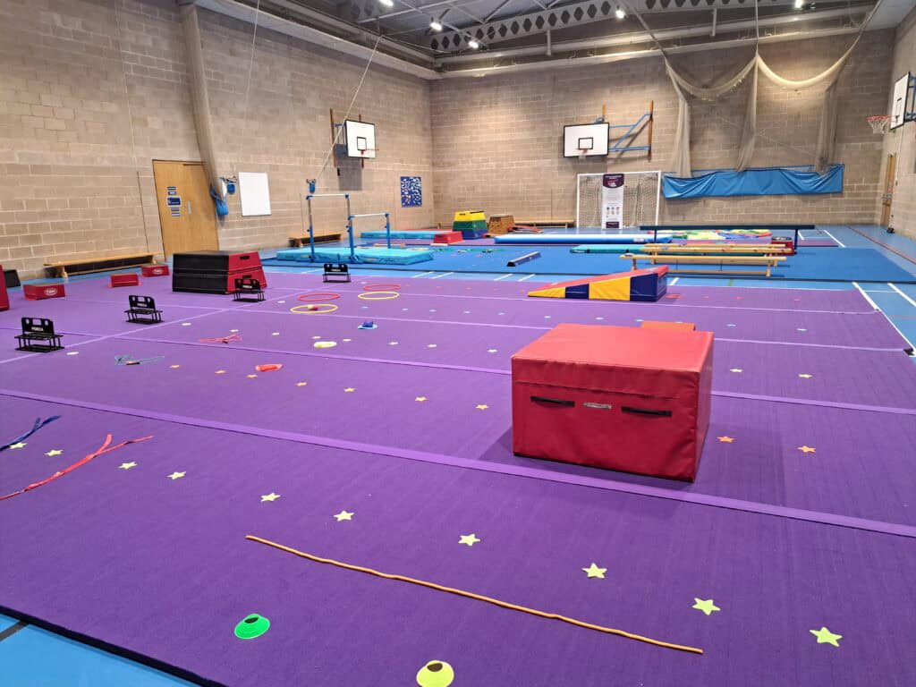 Jumping gymnastics exercises in a spacious indoor sports hall with purple mats, various training equipment, and safety gear for children and athletes to practice on.