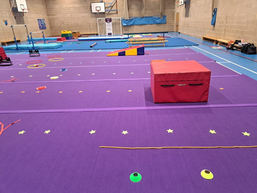 Climbing gym with padded purple flooring and various agility equipment for children and adults, including hurdles, rings, mats, and slacklines in a spacious indoor setting with brick walls and basketball hoops.