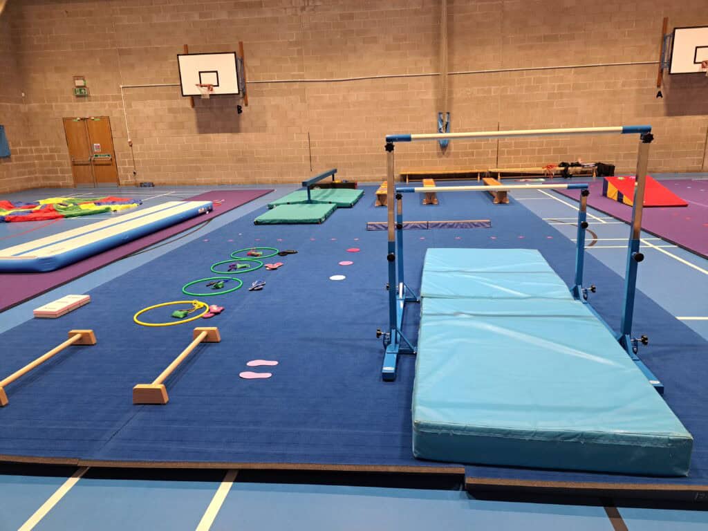 Brightly coloured gymnastics mats and equipment set up inside a gymnasium, including a vaulting table, trampolines, and balance beams, ready for gymnastics training or a competition.