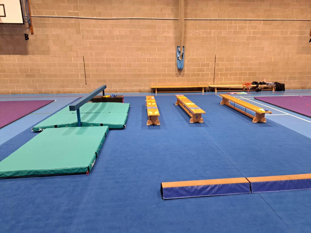 Climbing gym training area with crash mats, benches, and colourful gym equipment on a blue floor inside a sports centre. Suitable for gymnastics, training, and safety practice sessions.