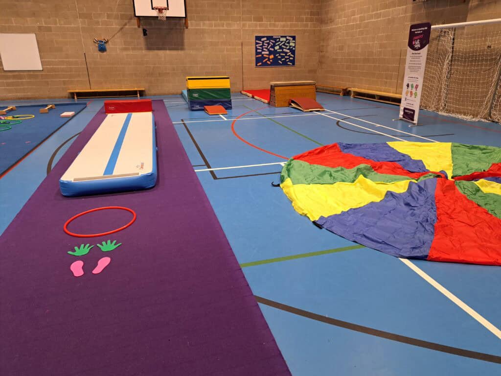 Fungymnastics obstacle course setup with colourful mats, hoops, and a parachute in an indoor sports hall. Suitable for children's physical activities and agility training.