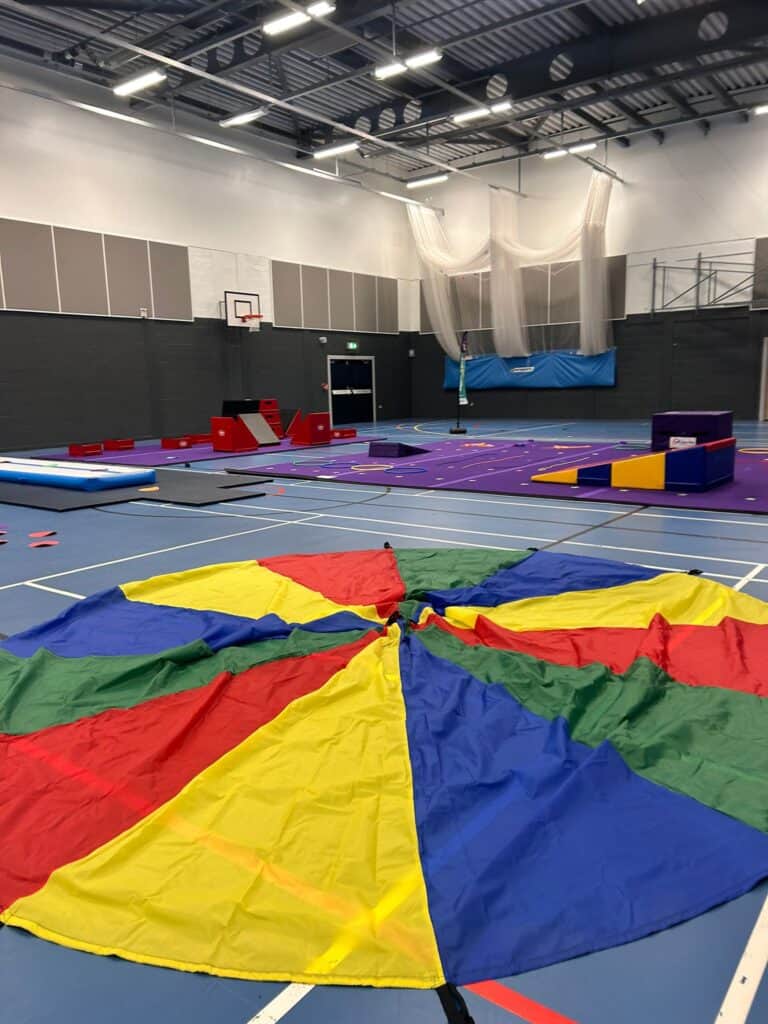 Active indoor gym with various equipment for physical activities, padded mats, colourful parachutes on the blue floor, high ceilings with ceiling lights, and protective netting for sports safety.