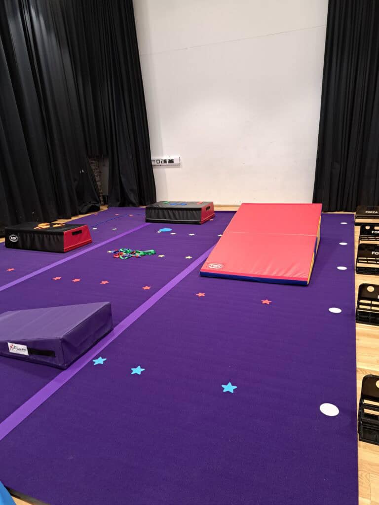 Indoor gymnastics training area with mats, foam blocks, and star-shaped floor markers on a purple mat, set against black curtains and a white wall.